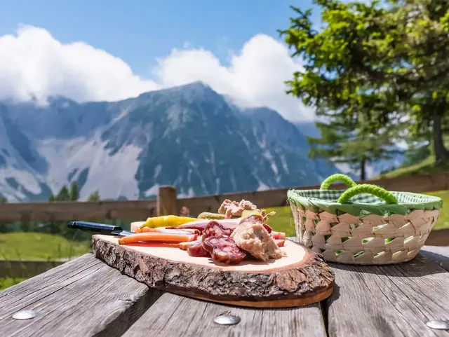 Traditionelle Brettljause mit Käse, Speck und Brot auf einem Holztisch, im Hintergrund ein weites Bergpanorama bei klarem Wetter.