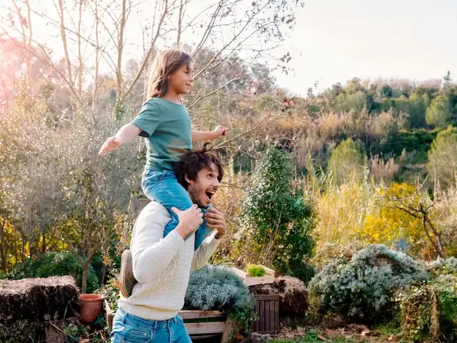 Vater trägt Sohn im Garten – Urlaub auf dem Bauernhof in der Holsteinischen Schweiz  - © tunedin – stock.adobe.com Ein Vater trägt seinen Sohn lachend auf den Schultern durch den Garten während eines Urlaubs auf dem Bauernhof in der Holsteinischen Schweiz.