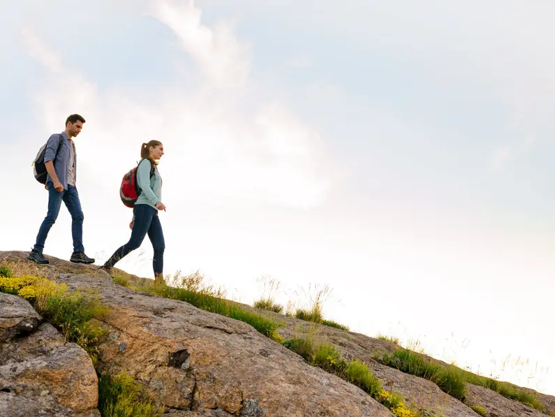 Couple of Young Happy Travelers Hiking with Backpacks on the Beautiful Rocky Trail at Warm Sunny Evening. Family Travel and Adventure Concept.