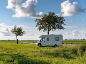 Ein weißer Campervan steht inmitten einer lebendigen Landschaft, eingerahmt von zwei Bäumen unter einem Himmel mit flauschigen Wolken – das Bild strahlt ein Gefühl von Freiheit aus.