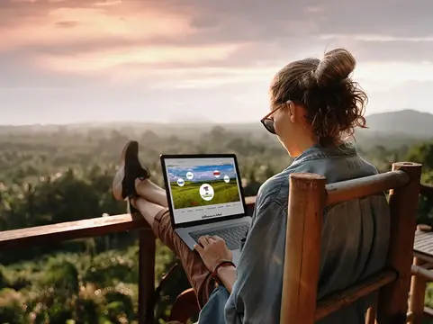 Ical Schnittstelle alt="Frau auf Terasse mit Blick über Wald arbeitet an Laptop. Auf dem Bildschirm sieht man symbolisch die Anbindemöglichkeiten mit Ical. "