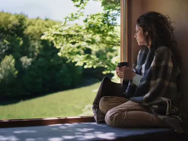 Frau sitzt am Fenster einer ländlichen Unterkunft und blickt in die grüne Landschaft