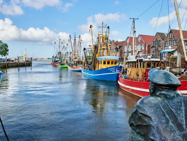 Metallfigur eines Matrosen blickt auf bunte Fischerboote im Hafen.