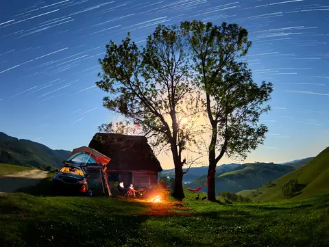 Ein Auto mit Dachzelt steht zwischen zwei großen Bäumen, daneben ein kleines, gemütlich brennendes Lagerfeuer. Über der Szene spannt sich ein klarer Nachthimmel voller funkelnder Sterne. 