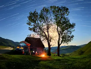 Ein Auto mit Dachzelt steht zwischen zwei großen Bäumen, daneben ein kleines, gemütlich brennendes Lagerfeuer. Über der Szene spannt sich ein klarer Nachthimmel voller funkelnder Sterne. 