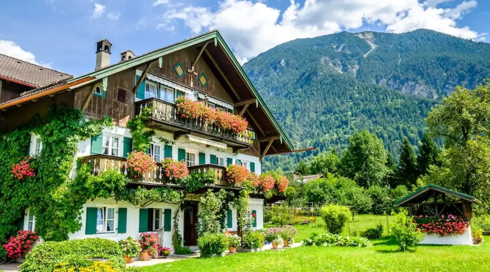 Bauernhaus mit vielen Blumen an den Balkonen und einem bewaldeten Berg im Hintergrund - eine Ferienwohnung in Bayern auf dem Land