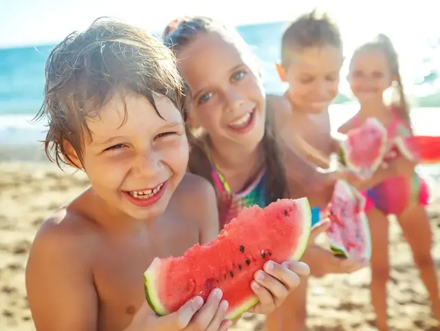 Fröhliche Strandmomente – Ferienwohnung auf Fehmarn Vier Kinder in Badesachen stehen am Strand vor dem Meer und essen gemeinsam Melone auf Fehmarn.
