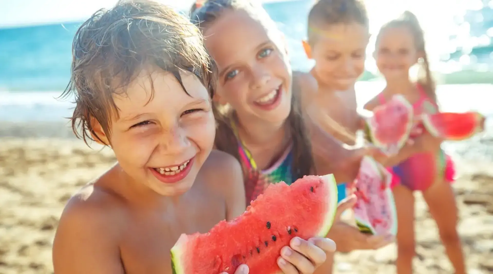 Vier Kinder in Badesachen stehen am Strand vor dem Meer und essen gemeinsam Melone auf Fehmarn.