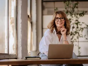 Einfach mehr Buchungen - mit Landsichten Frau sitzt in Küche vor Laptop und lächelt in die Kamera.