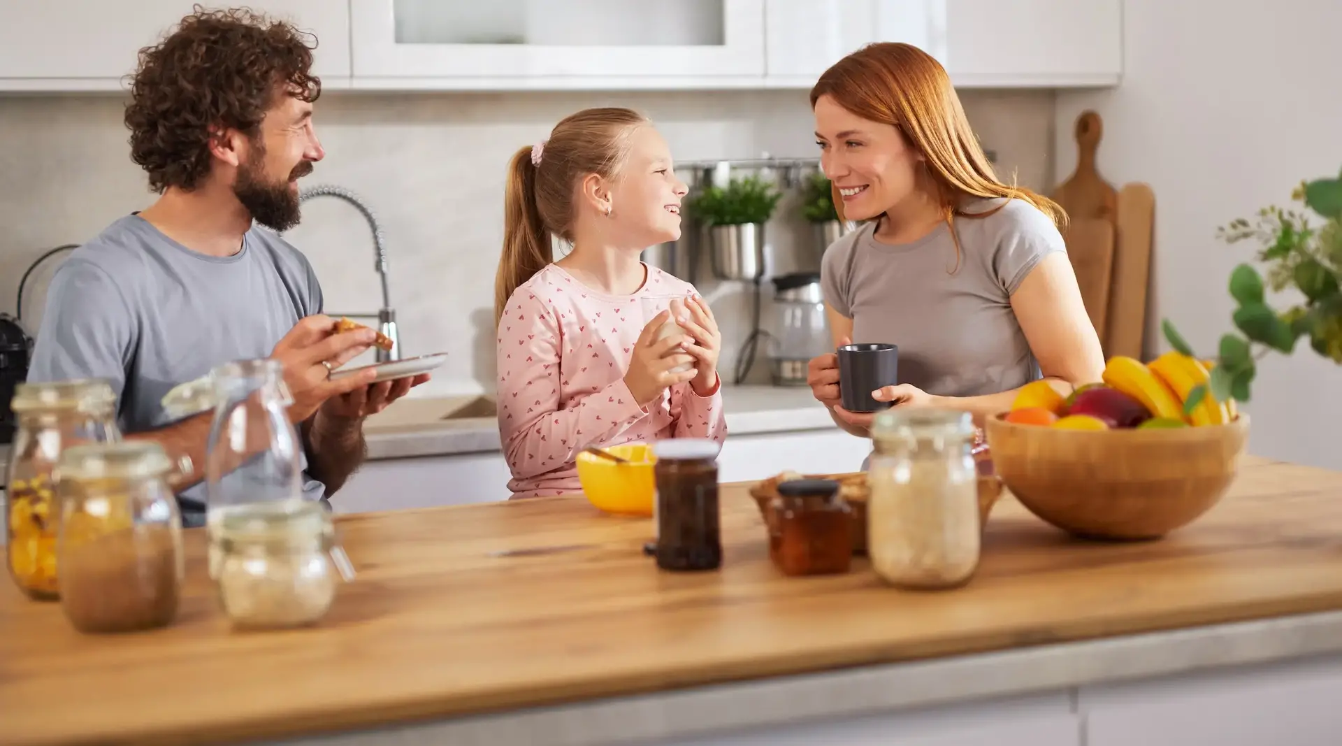 Eine Familie mit Mutter, Vater und Tochter sitzt fröhlich am Frühstückstisch in einer Ferienwohnung in Bayern. 