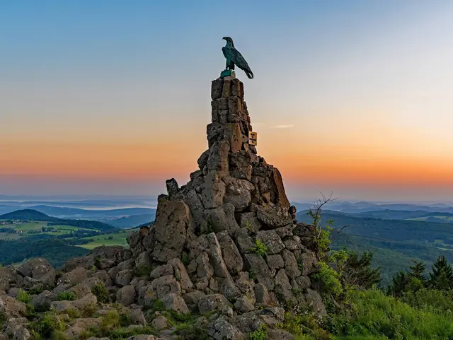 Fliegerdenkmal auf der Wasserkuppe in der Rhön mit steinernem Sockel und Adlerfigur vor dem orangefarbenen Morgenhimmel.