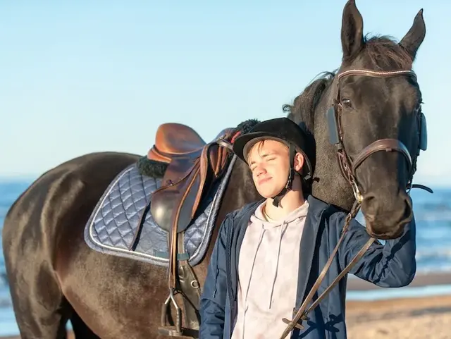 Junger Mann mit Reiterhelm steht zusammen mit Pferd am Strand. 
