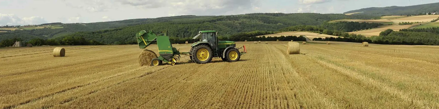 Traktor mit Ballenpresse fährt übers Stoppelfeld und Presst runde Strohballen. 