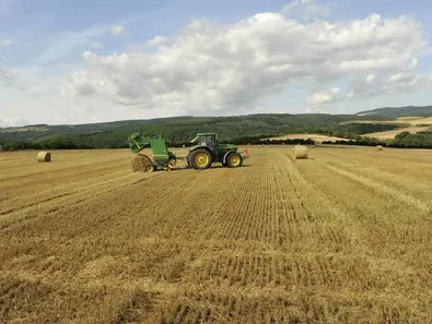 Traktor mit Ballenpresse fährt übers Stoppelfeld und Presst runde Strohballen. 