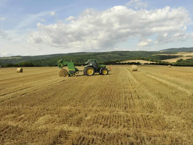 Traktor mit Ballenpresse fährt übers Stoppelfeld und Presst runde Strohballen. 