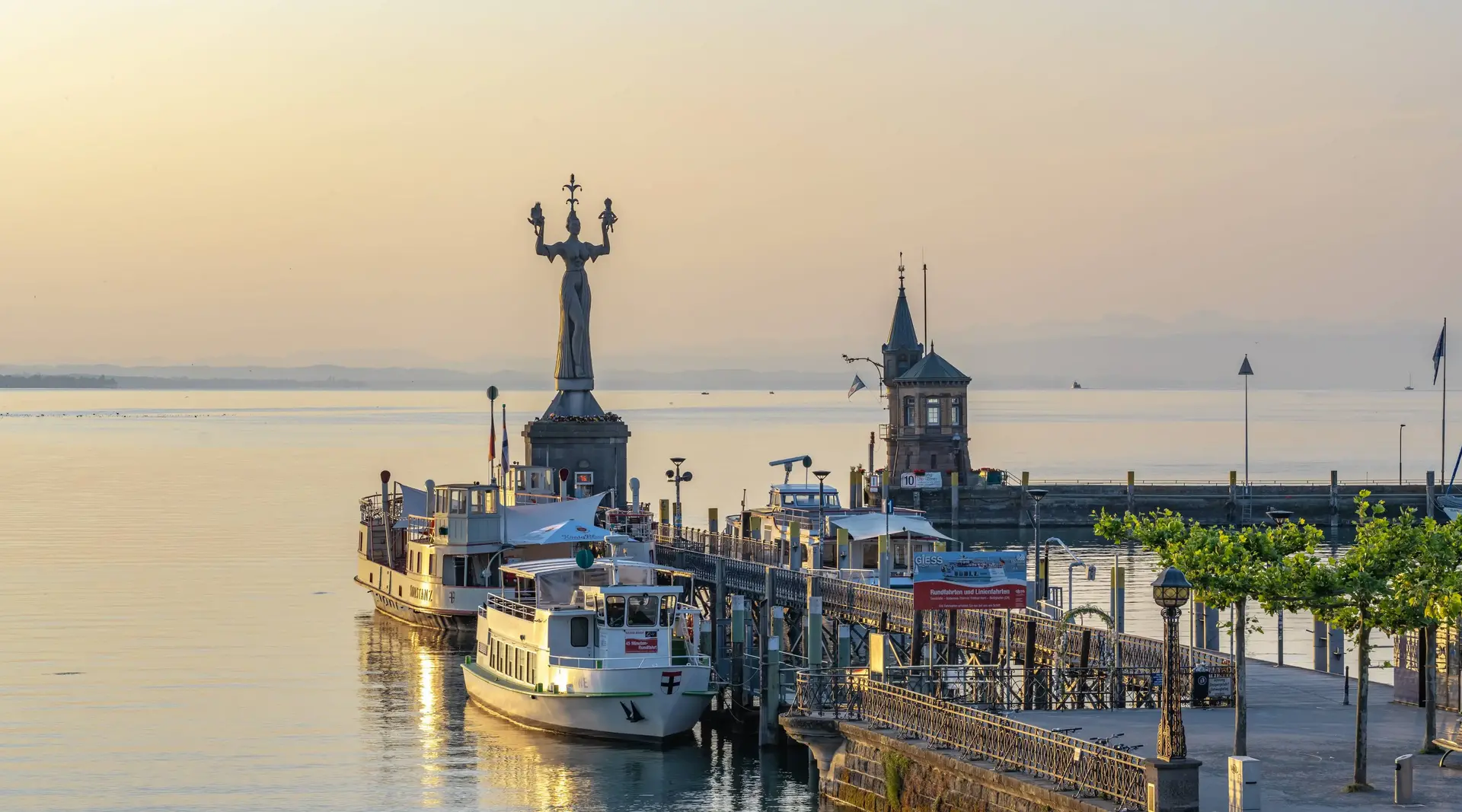 Konstanz am Bodensee – Hafeneinfahrt bei Sonnenaufgang - © Michael, Adobe Stock Hafeneinfahrt von Konstanz mit Imperia-Statue, Katamaran auf dem Bodensee und Landschaftspanorama bei Sonnenaufgang.