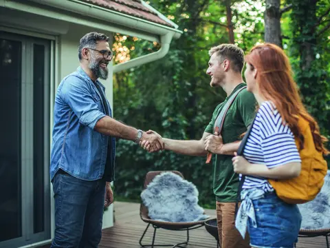 1017057100 alt="Young couple renting house. House owner shaking hands with couple who rent his house. Couple is on vacation and they are wearing backpacks."