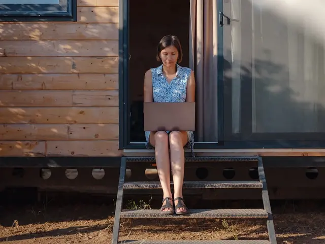 Frau sitzt barfuß vor dem Eingang eines Tiny Houses aus Holz in ländlicher Umgebung