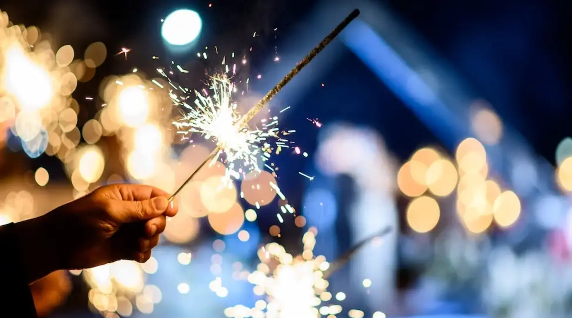 Hand hält eine brennende Wunderkerze vor festlich beleuchtetem Hintergrund bei einer Silvesterfeier im Winter