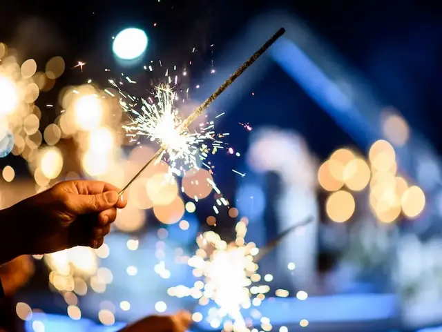 Hand hält eine brennende Wunderkerze vor festlich beleuchtetem Hintergrund bei einer Silvesterfeier im Winter
