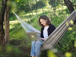 Frau in Hängematte in grünem Garten lächelt in Laptop hinein. 