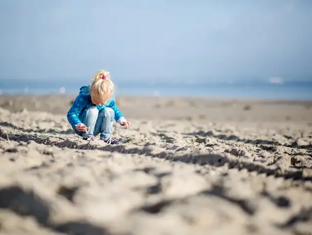 Buddelspaß am Strand Mädchen mit blonden Haaren spielt mit den Händen im Sand. Im Hintergrund sieht man das Meer und blauen Himmel.