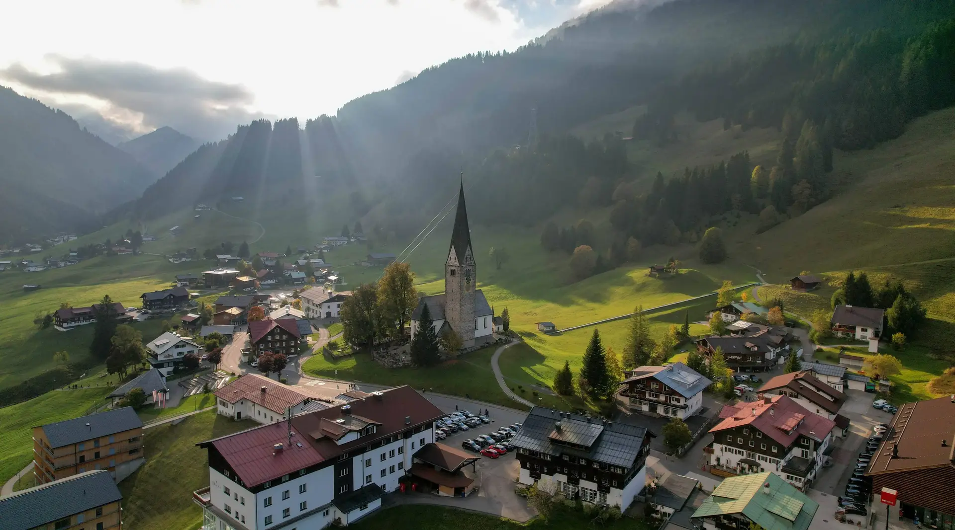 Luftaufnahme des Bergdorfs Mittelberg im Kleinwalsertal mit Häusern, grünen Wiesen und umliegenden Alpengipfeln.