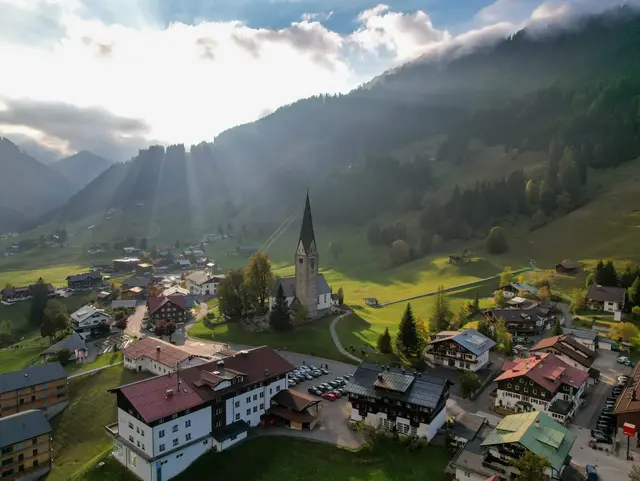 Luftaufnahme des Bergdorfs Mittelberg im Kleinwalsertal mit Häusern, grünen Wiesen und umliegenden Alpengipfeln.