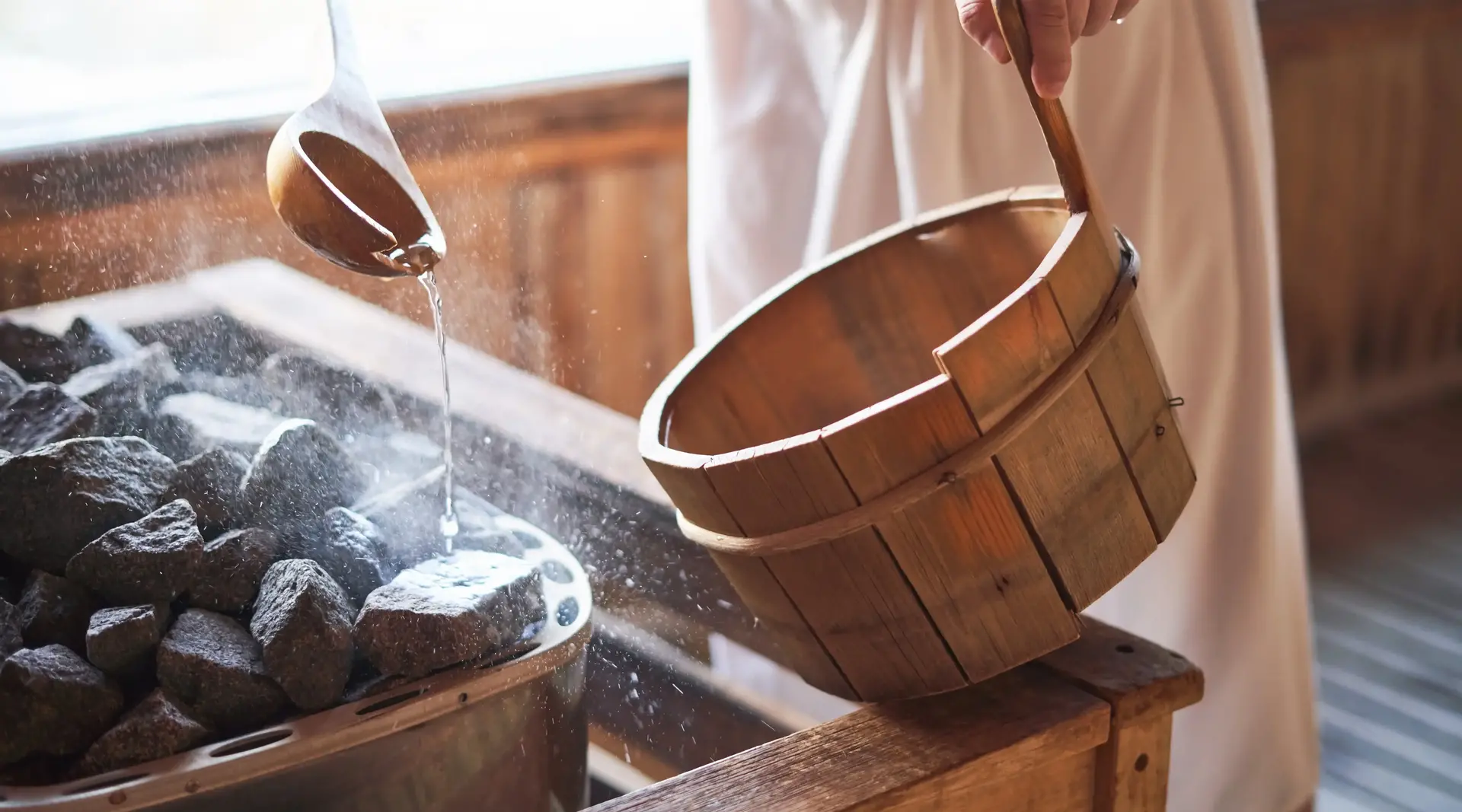 Aus einem Holzeimer wird in einer Sauna mit Holzlöfel Wasser auf heiße steine geleert.