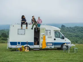 Auf einer grünen Wiese, vor dem Hintergrund eines Waldes, sitzen drei junge Menschen entspannt auf dem Dach eines weißen Campervans – ein Moment voller Freiheit und Naturverbundenheit.
