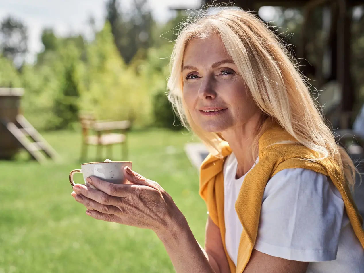 Cheerful woman enjoying hot drink among nature Relaxed smiling mature lady is holding cup of coffee while having rest in yard of countryside house