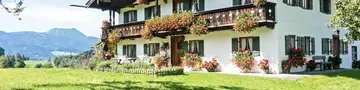 Traditioneller bayerischer Bauernhof / Ferienhof mit Holzfassade und Balkonen, liebevoll mit Blumen geschmückt. Große Wiese im Vordergrund, Alpenpanorama im Hintergrund, sonnige Wetterlage. Eine perfekte Ferienwohnung in Bayern auf dem Land. 