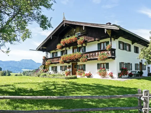 Traditioneller bayerischer Bauernhof / Ferienhof mit Holzfassade und Balkonen, liebevoll mit Blumen geschmückt. Große Wiese im Vordergrund, Alpenpanorama im Hintergrund, sonnige Wetterlage. Eine perfekte Ferienwohnung in Bayern auf dem Land. 