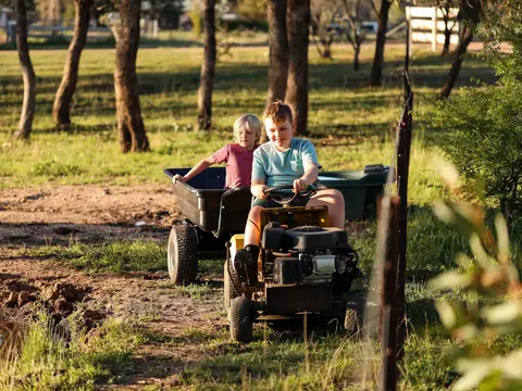 Kinder beim Spielen auf einem Erlebnishof - © Caseyjadew, AdobeStock alt="Zwei Jungs die glücklich auf einem Tretttraktor fahren. Der zweite Junge lässt sich in einem Anhänger durch die Natur ziehen."