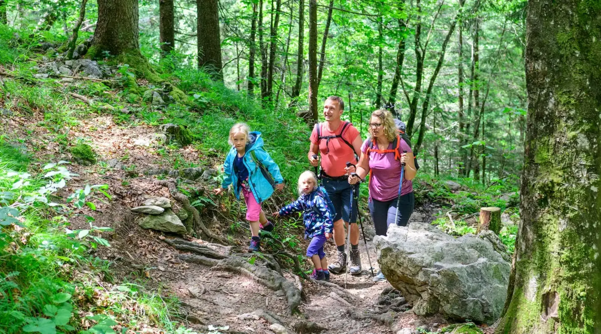 Eine Familie mit zwei Mädchen, Mutter und Vater wandert bei warmen Temperaturen bergauf durch einen Wald. 