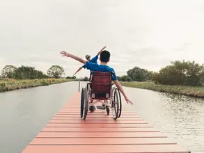 Mann im Rollstuhl sitzt mit seinem Vater auf einem Holzsteg an einem See während eines barrierefreien Urlaubs auf dem Land