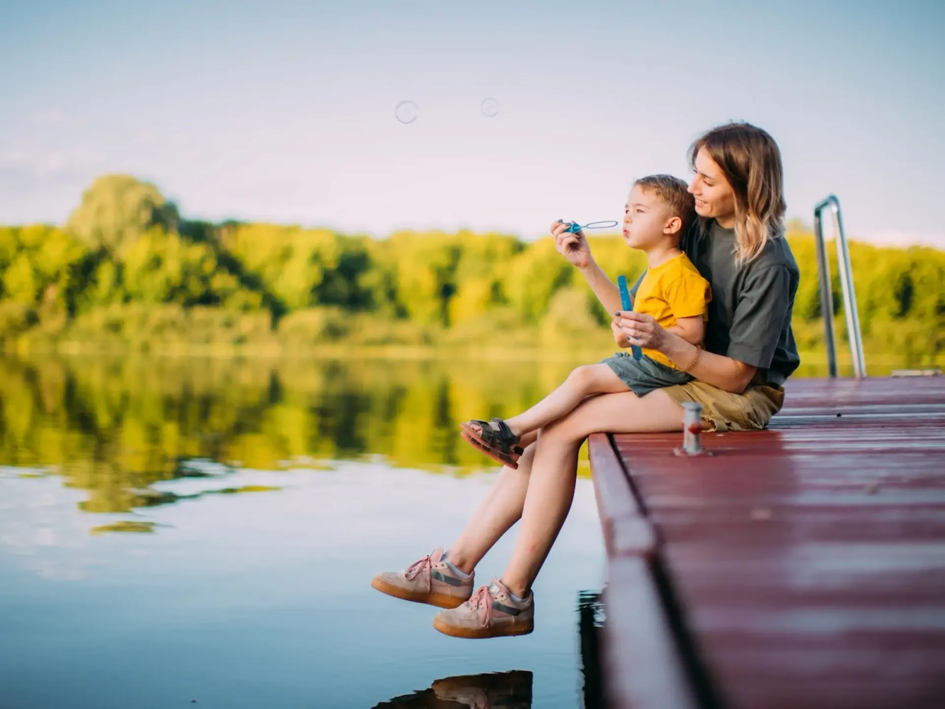 Mutter und kleiner Junge sitzen auf einem Holzsteg in ländlicher Natur und lassen Seifenblasen steigen – idyllische Szene im Sommerurlaub beim Ferienhaus auf dem Land. 