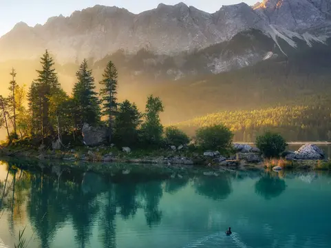 Idyllischer See bei Sonnenaufgang - © Smileus – stock.adobe.com alt="Idyllischer See bei Sonnenaufgang, ein malerisches Panorama mit majestätischen Bergen und dem goldenen Sonnenlicht, das hinter den Bäumen hereinscheint"