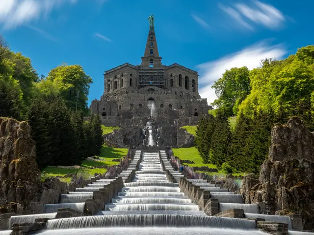Monumentale Wasserkaskaden im Bergpark Wilhelmshöhe in Kassel mit dem Herkules-Denkmal im Hintergrund, umgeben von grünen Bäumen.