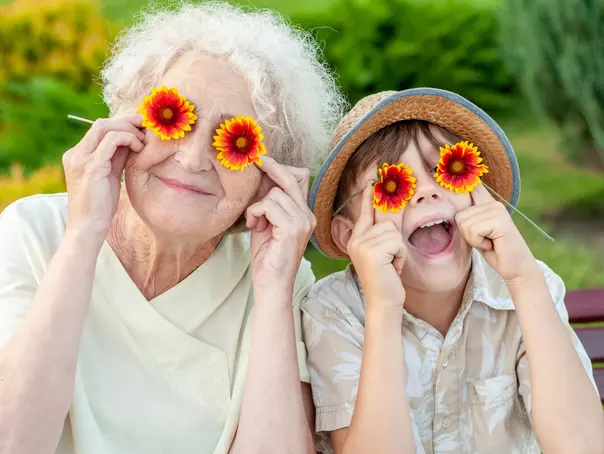 Boy and grandmother puts on the eyes of chamomile. Family holiday.Laughing grandson with his beloved grandmother spend leisure time outdoors. An elderly woman and a grandchild. Summer spring time