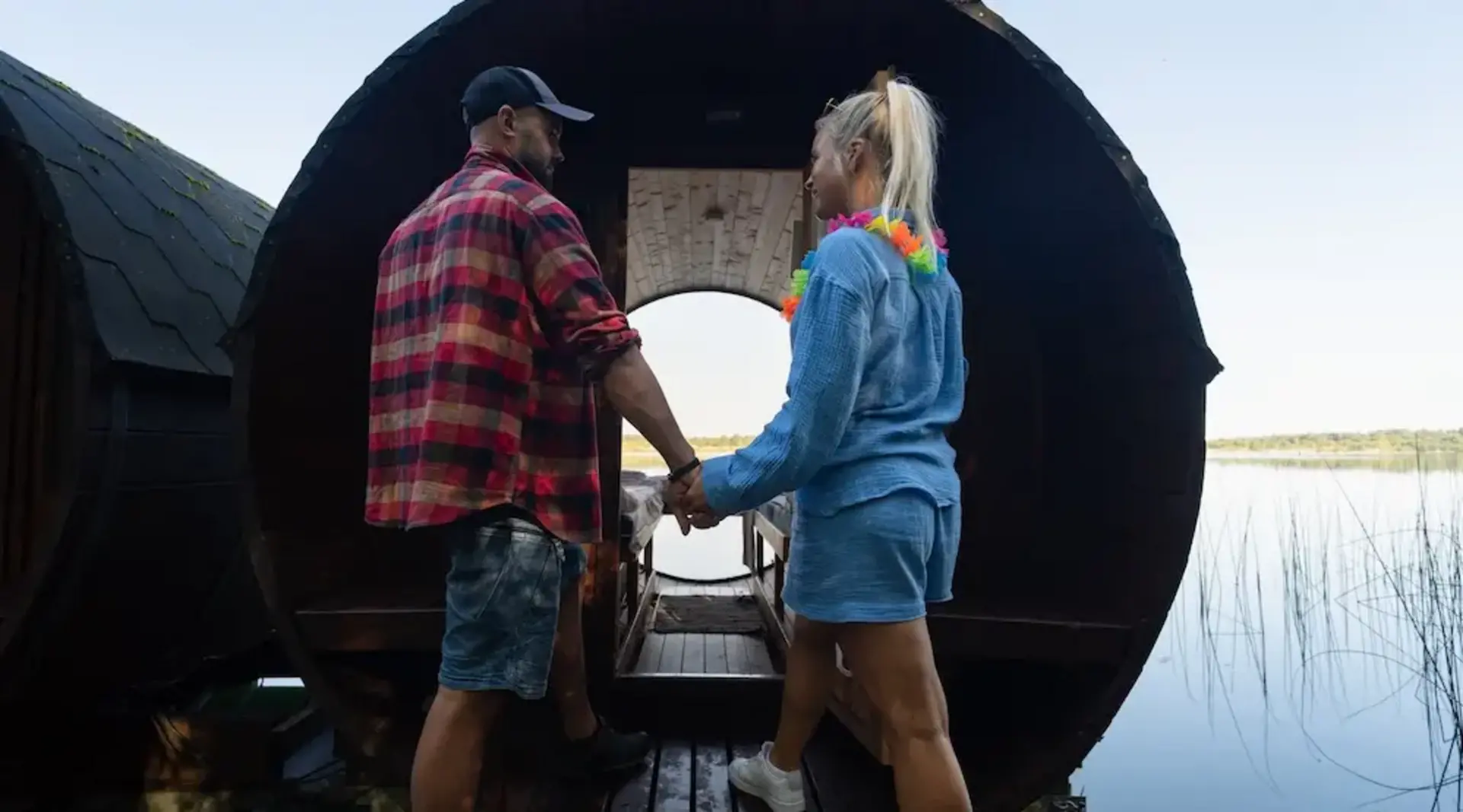 Zwei Personen stehen vor einem Holz-Weinfass am See und gehen gemeinsam rein