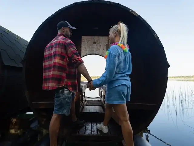 Zwei Personen stehen vor einem Holz-Weinfass am See und gehen gemeinsam rein