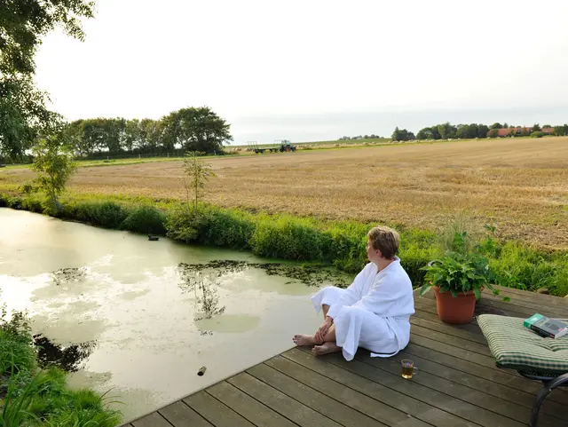 Frau sitzt im Bademantel auf Steg am Fluss