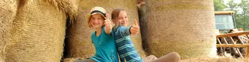 Zwei glückliche Mädchen sitzen auf Heuballen in der Scheune und halten ihre Daumen nach oben
