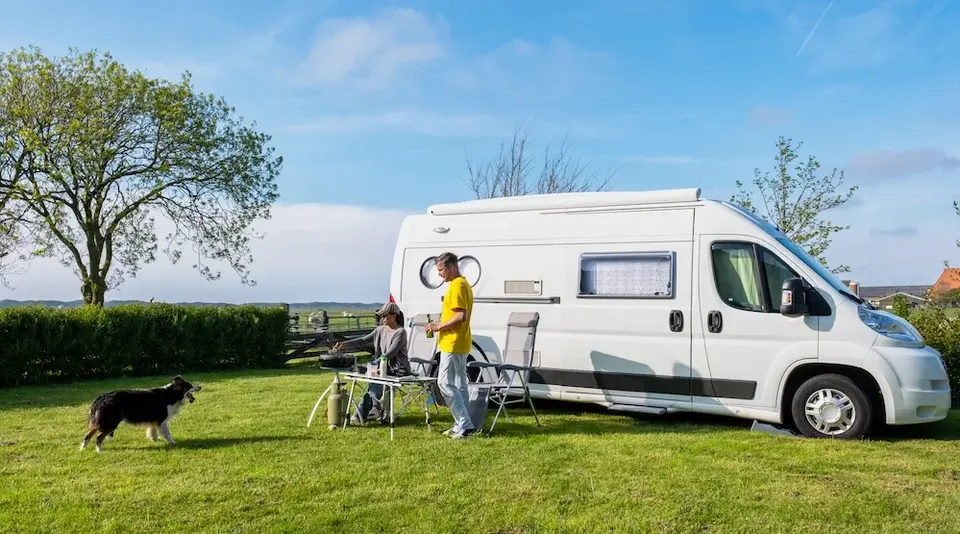 Zwei Menschen und ihr Hund genießen die Natur vor ihrem weißen Camper-Kastenwagen – Campingidylle auf der Wiese. 