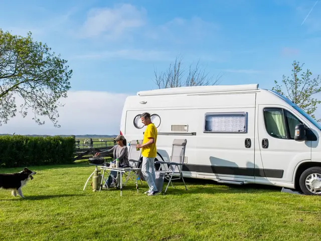 A man and his faithful dog stand next to an RV or camper van in a picturesque grassy field in Texel Netherlands, soaking in the serenity of their surroundings. a couple at a camping farm - © Fokke Baarssen, adobe stock Zwei Menschen und ihr Hund genießen die Natur vor ihrem weißen Camper-Kastenwagen – Campingidylle auf der Wiese.