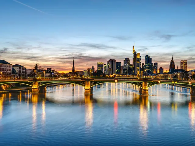Beleuchtete Skyline von Frankfurt am Main in der Abenddämmerung mit Hochhäusern und Spiegelungen auf dem Main.