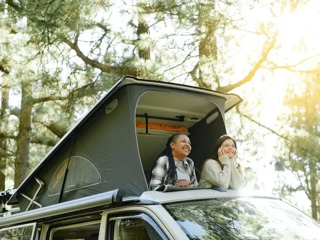 Zwei Frauen schauen aus dem geöffneten Klappdach einen Campingbuses in die von Bäumen bewachsene Umgebung.