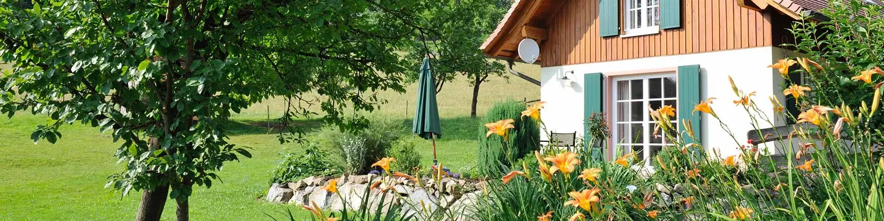 Kleines Haus mit grünen Fensterläden und Holzschirm umgeben von Natur und gelben Lilien