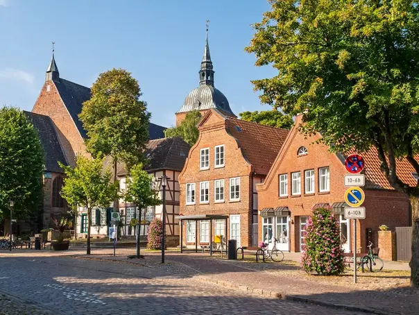 Eine Backstein-Häuserreihe mit Kirche ist von grünen Bäumen umgeben und unter blauem Himmel auf Fehmarn zu sehen.